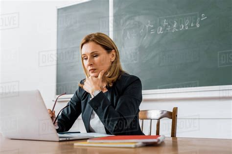 Female Teacher Sitting At Desk And Using Computer During Lesson In