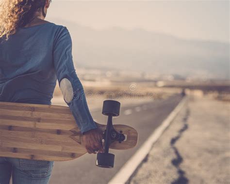 Carefree Woman Standing And Holding Wooden Longboard Behind Her Back On