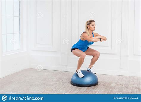 woman in black shorts and blue top working in gym doing exersice in bosu balance trainer squats