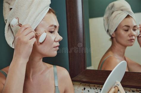 Close Up Woman Using Jade Facial Roller For Face Massage Sitting In Bathroom Looking In The