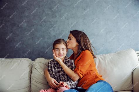 Счастливая мать обнимает и целует свою любимую дочь сидя на диване концепция счастливой семьи