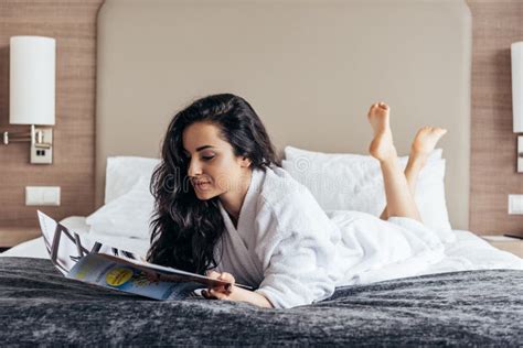 Barefoot Brunette Young Woman In White Bathrobe Lying On Bed And Reading Magazine Stock Image
