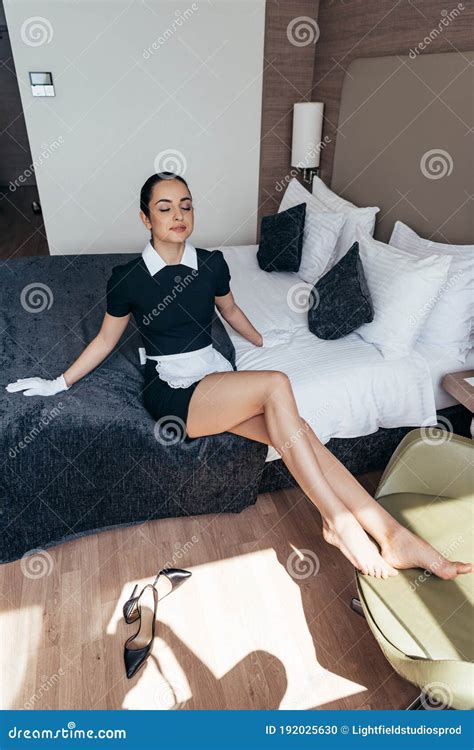 Maid In White Apron And Gloves Sitting On Bed With Closed Eyes In Hotel Room Stock Photo Image