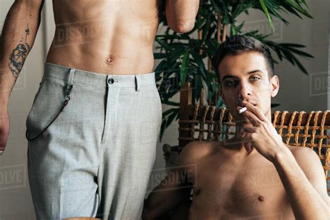 stylish shirtless gay couple posing in front of the camera sitting in a chair smoking cigarette
