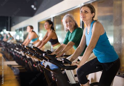 Positive Mature Female Riding Exercise Bike During Cycling Class In Modern Gym Stock Photo