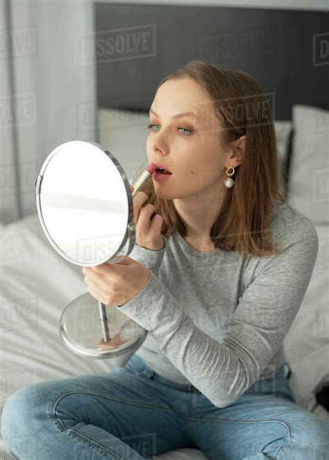 Woman Sitting On Bed Putting Lipstick On Her Lips In Front Of The