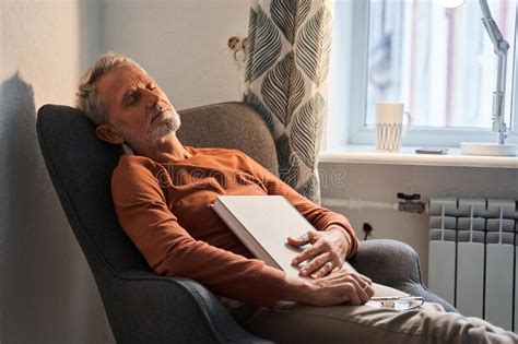 Tired Mature Man Sleeping At The Armchair With Book On Chest After