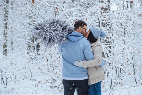 A Loving Couple In The Winter Forest Is Kissing A Man Holds A Christmas Tree On His Shoulder