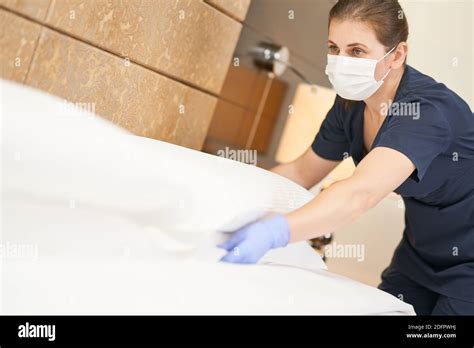 Maid In Mask Fluffing White Pillow On Bed While Cleaning The Room In
