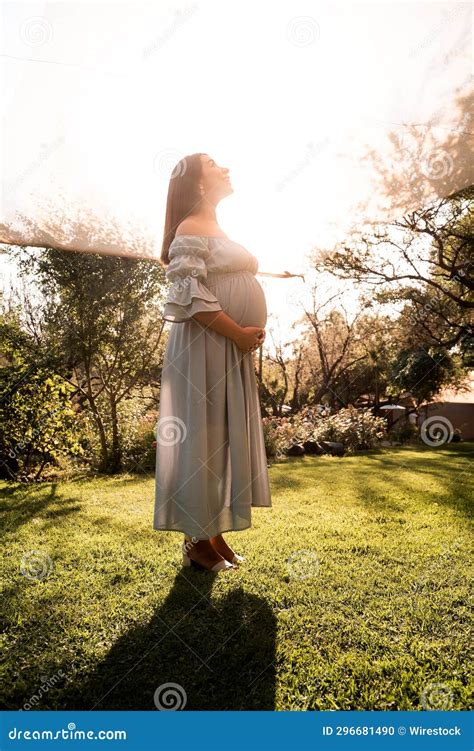 pregnant latina woman in a dress standing in lush green sunlit park hands gently cradling her