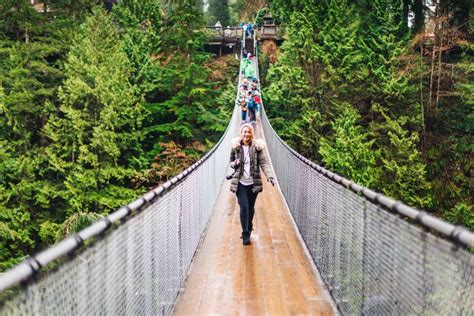 a woman walking across a suspension bridge over a forest filled with lots of tall trees