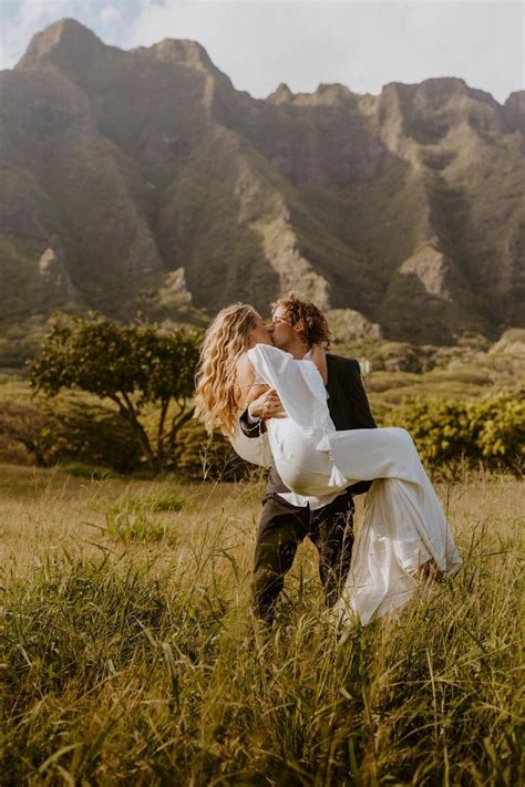 A Bride Carrying Her Groom Through The Tall Grass In Front Of Some Mountain Range Behind Them