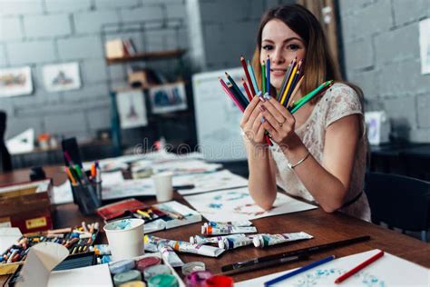 Smiling Young Woman Holding A Bunch Of Crayons Sitting At Desk