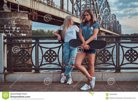 Two Beautiful Hipster Girls Standing With Skateboard Against A Bridge