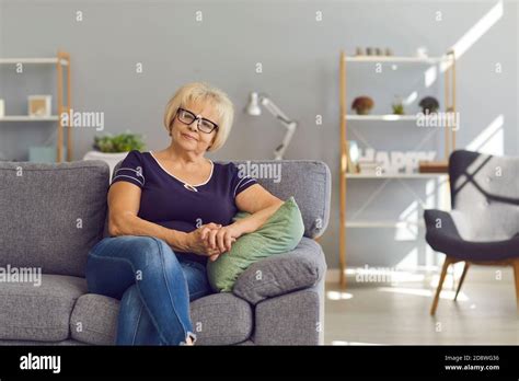 Mature Woman Sitting On Comfortable Sofa In Her Modern Living Room And Looking At Camera Stock