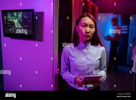 Portrait Of Serious Asian Female Network Administrator Standing With Tablet Against Computer