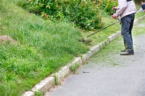 A Utility Worker Mows Tall Green Grass With A Petrol Trimmer Mower