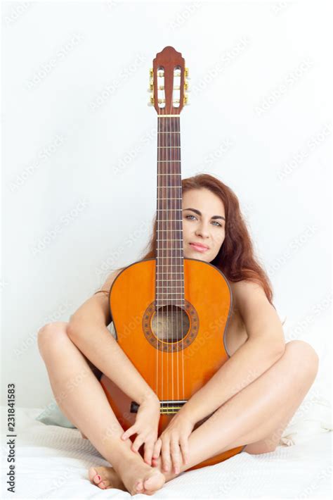 Happy Young Nude Woman Sitting On Her Bed And Hugging An Acoustic