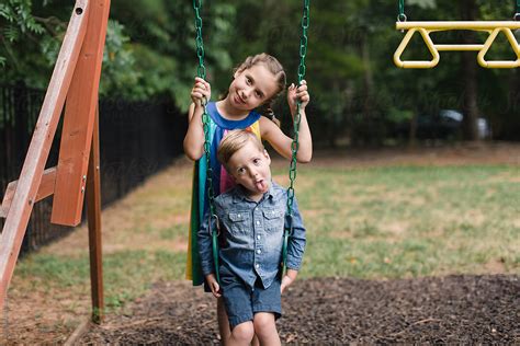 Big Sister Standing Behind Younger Brother Who Is Sitting On A Swing By Stocksy Contributor