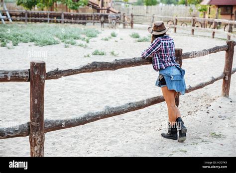 Back View Of Young Woman Cowgirl In Hat Standing Near Fence On Ranch