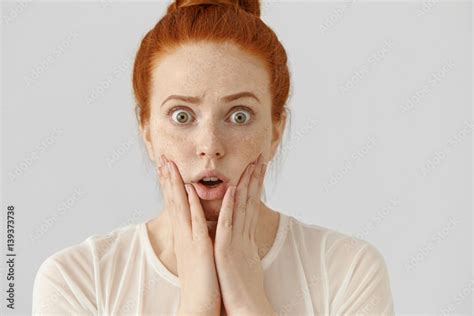 Omg Headshot Of Funny Bug Eyed Girl With Ginger Hair Keeping Hands On Cheeks Having Shocked Or
