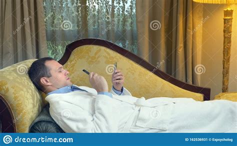 Businessman In A Bathrobe Relaxes On A Sofa With A Phone And Smokes A