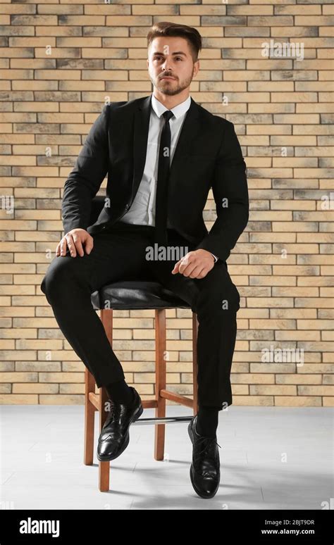 Handsome Man In Formal Suit Sitting On Chair Against Brick Wall Stock