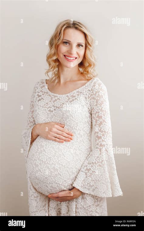 Portrait Of Smiling White Caucasian Blonde Pregnant Woman In White Lacy Dress Touching Her Belly