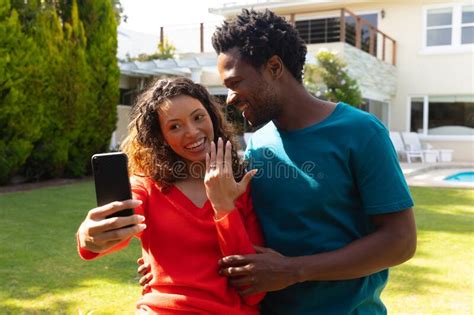 Smiling Young Multiracial Couple Showing Engagement Ring While Taking Selfie On Smartphone In