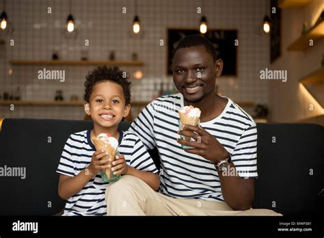 Happy African American Father And Son Eating Ice Cream And Smiling At Camera While Sitting