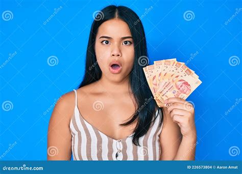 Young Beautiful Asian Girl Holding Mexican Pesos Scared And Amazed With