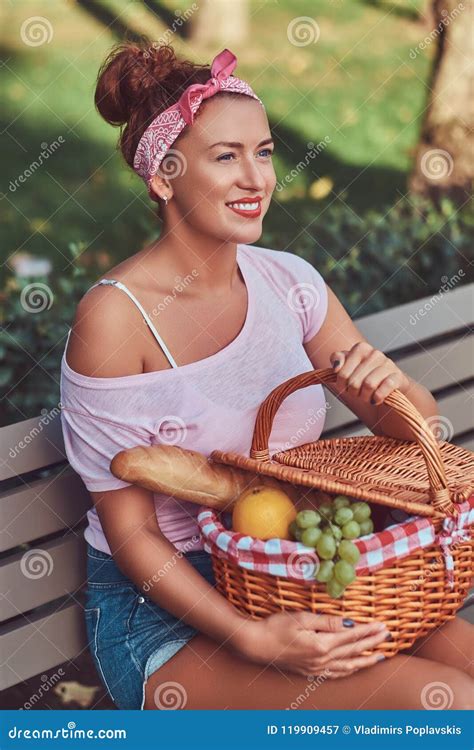 Happy Beautiful Redhead Female Wearing Casual Clothes Holds A Picnic Basket While Sitting On A