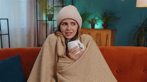 Sick Young Woman Wear Hat Wrapped In Plaid Sit Alone Shivering From Cold On Sofa Drinking Hot