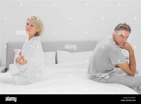 Couple Sitting On Opposite Sides Of Bed Looking At Camera After A Fight
