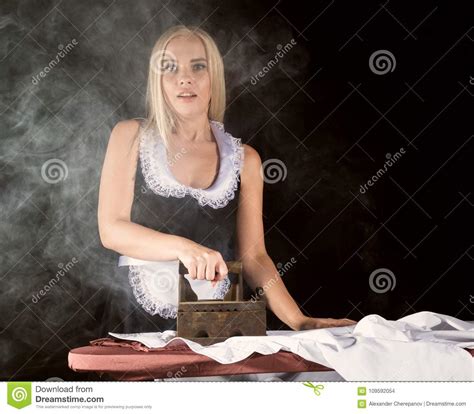 Blonde Woman In Housekeeper Suit Ironing White Shirt With Old Iron Retro Style On A Dark