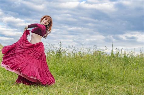 Young Red Haired Girl In Red Dance Dress Dances On Green Meadow Against