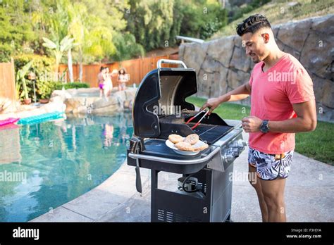 Young Man Cooking Burgers On Barbecue Next To Garden Swimming Pool