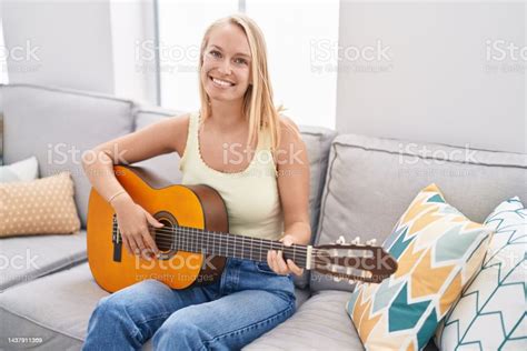 Young Blonde Woman Playing Classical Guitar Sitting On Sofa At Home