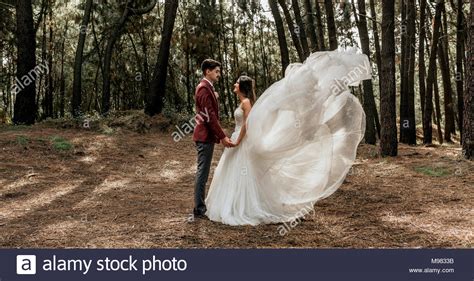 Bride With Windswept Wedding Dress And Groom Standing In Forest Holding