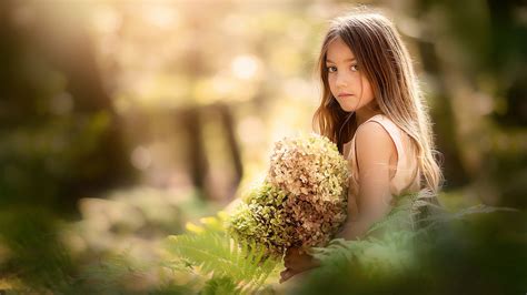 Cute Little Girl Is Standing Near Green Plant With Flower Bouquet