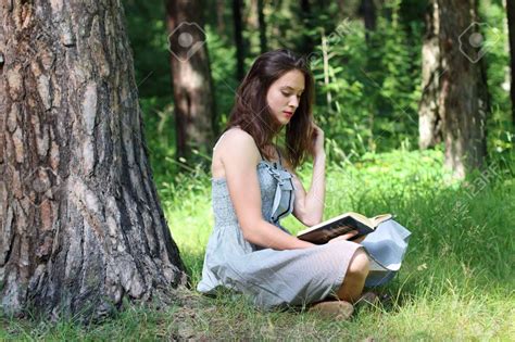 Beautiful Girl In Dress Sitting Under Tree On Grass And Reading Book