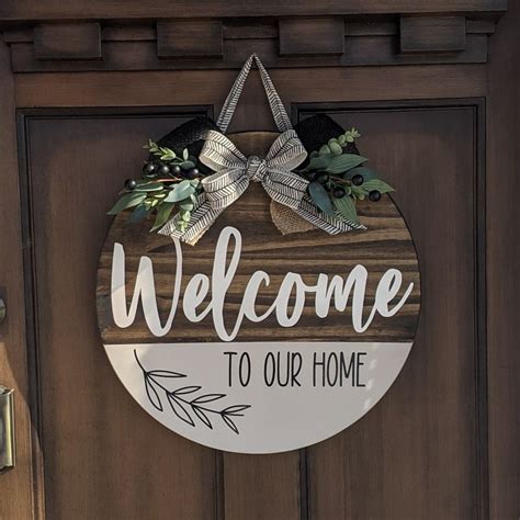 A Welcome To Our Home Sign Hanging On The Front Door With Greenery And Bow