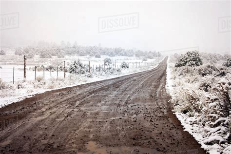 Wet Dirt Road Amidst Snow Covered Field During Foggy Weather Stock