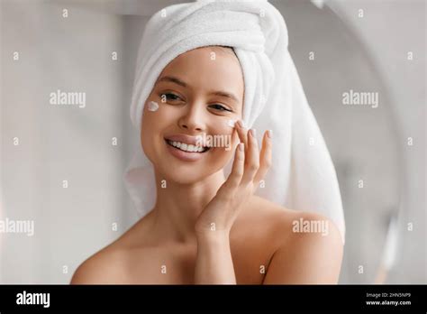 Headshot Of Woman Applying Facial Cream On Cheeks Smiling At Her Reflection In Mirror Enjoying