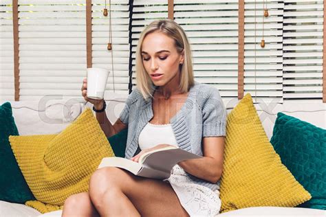 Beautiful Woman Relaxing And Reading A Book And Drinking Coffee Sitting On Sofa In Her Living