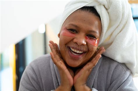 Happy Plus Size African American Woman Wearing Robe Applying Eye Masks In Bathroom Stock Image