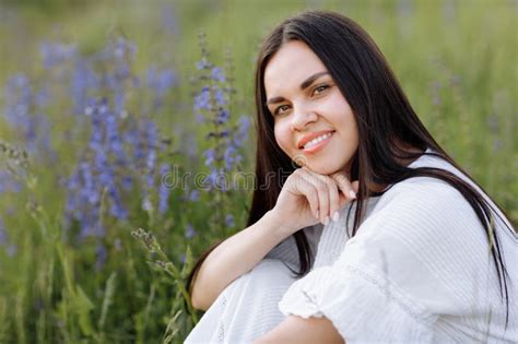 Young Smiling Beautiful Brunette Woman In White Dress Is Sitting In Green Meadow With Lupine