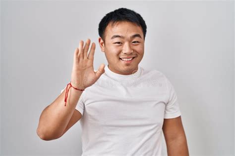 Young Chinese Man Standing Over White Background Waiving Saying Hello