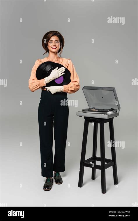 happy stylish woman in vintage clothes holding vinyl discs while posing near record player on