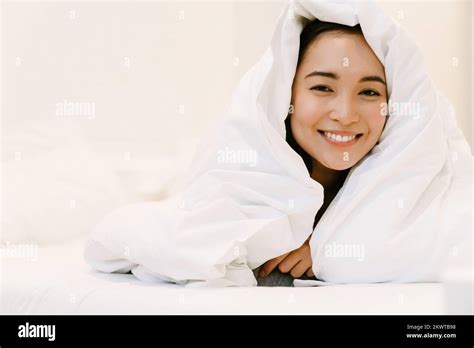 Young Smiling Asian Girl In Home Suit Lying On Bed Under Covers At Home